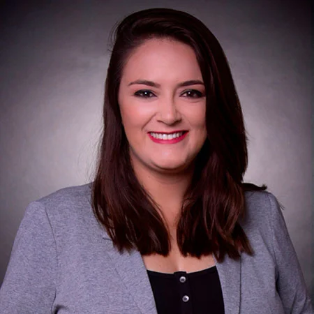 Retrato de uma mulher de pele clara com cabelos escuros abaixo dos ombros, sorrindo, de terno cinza.