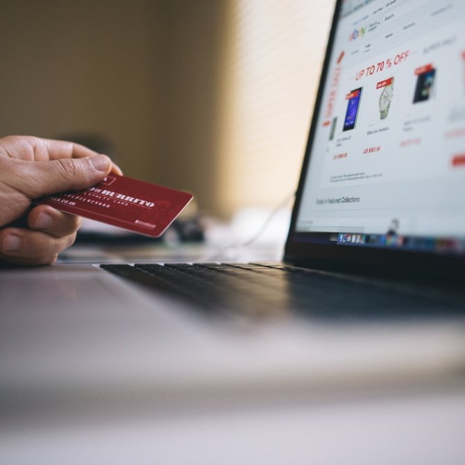 Imagem de um notebook aberto e uma mão de pele clara segurando um cartão de crédito vermelho indicando um processo de compra online.