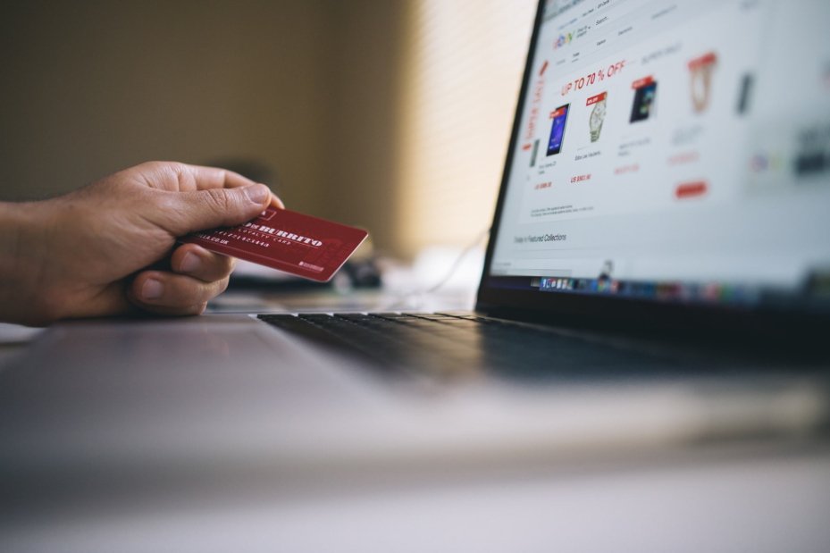 Imagem de um notebook aberto e uma mão de pele clara segurando um cartão de crédito vermelho indicando um processo de compra online.